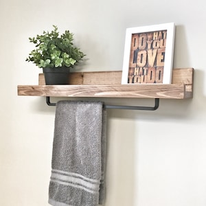 Op de afbeelding: Een rustiek houten plank met een zwarte handdoekstang, met een grijze handdoek. Een kleine potplant en een ingelijste print met de tekst "DO WHAT YOU LOVE" staan op de plank. De plank is aan een lichtgekleurde muur bevestigd.
