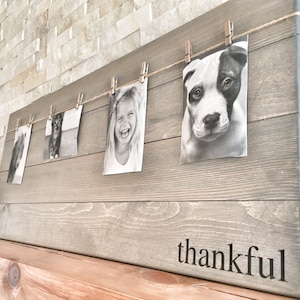 May include: A rustic wooden photo display with a rope and pegs holding black and white photos of a cat, a girl, and a dog. The wood is grey and has the word "thankful" written on the bottom.