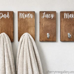 May include: Four wooden wall hooks with white text that reads "Dad", "Mom", "Lucy", and "Max". Two hooks have towels hanging from them. The wooden planks are a medium brown color. The background is a light gray.