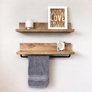 Peut inclure: Deux étagères flottantes en bois avec une finition naturelle. L'étagère supérieure contient une bougie et une impression encadrée qui dit "DO WHAT YOU LOVE WHAT YOU DO". L'étagère inférieure a une barre porte-serviettes noire et une serviette grise. Une petite barre de savon est sur l'étagère inférieure.
