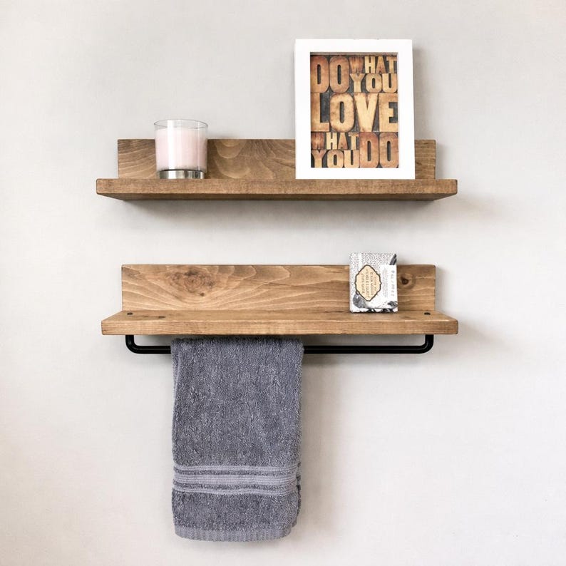 May include: Two wooden floating shelves with a natural finish. The top shelf holds a candle and a framed print that reads "DO WHAT YOU LOVE WHAT YOU DO." The bottom shelf has a black towel bar and a gray towel. A small soap bar sits on the bottom shelf.