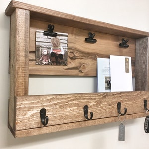 May include: Rustic wooden wall organizer with a shelf, hooks, and clips. The shelf has a photo and some papers on it. The hooks have keys and a key chain hanging from them.