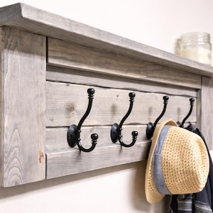 May include: A rustic wooden coat rack with a shelf and four black metal hooks. The rack is made of gray-toned wood and has a shelf on top. A straw hat with a blue band hangs from one of the hooks.