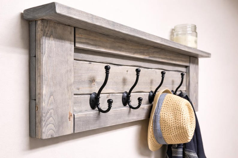 May include: A rustic wooden coat rack with a shelf and four black metal hooks. The rack is made of gray-toned wood and has a shelf on top. A straw hat with a blue band hangs from one of the hooks.