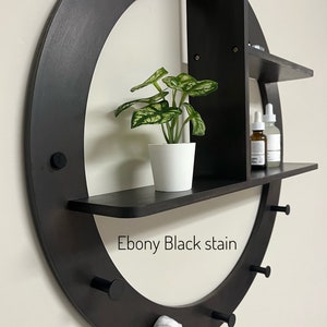 May include: A black circular wall shelf with three shelves and hooks. The shelf is stained ebony black and has a 24 inch diameter. The top shelf has a small potted plant, the middle shelf has two bottles, and the bottom shelf is empty. The hooks are on the bottom of the shelf and have a towel hanging on one of them. "Ebony Black stain" and "Shown in the 24 inch circle size" are written on the shelf.