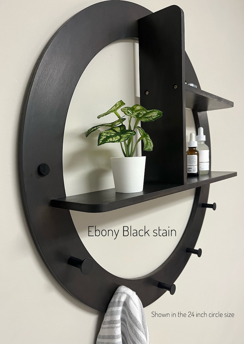 May include: A black circular wall shelf with three shelves and hooks. The shelf is stained ebony black and has a 24 inch diameter. The top shelf has a small potted plant, the middle shelf has two bottles, and the bottom shelf is empty. The hooks are on the bottom of the shelf and have a towel hanging on one of them. "Ebony Black stain" and "Shown in the 24 inch circle size" are written on the shelf.