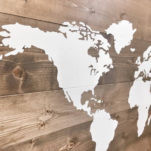 Pode incluir: Um mapa mundial pintado de branco sobre um fundo de madeira marrom. O mapa &eacute; delineado em branco e os continentes s&atilde;o preenchidos com tinta branca.