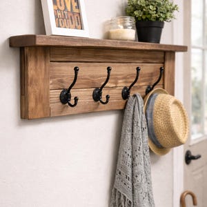 May include: A rustic wooden wall shelf with four black metal hooks. A framed print with the text "DO WHAT YOU LOVE THAT YOU DO" sits on the shelf, along with a potted plant, a candle, a gray scarf, and a straw hat.