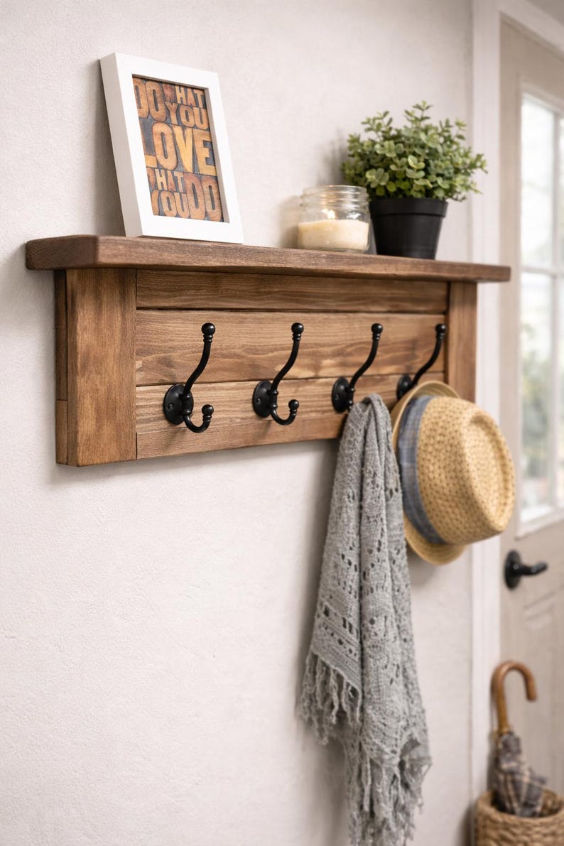May include: A rustic wooden wall shelf with four black metal hooks. A framed print with the text "DO WHAT YOU LOVE THAT YOU DO" sits on the shelf, along with a potted plant, a candle, a gray scarf, and a straw hat.