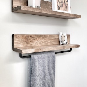 May include: Two wooden floating shelves with a light brown finish are mounted on a white wall. The top shelf holds a candle and a framed print with the text "DO WHAT YOU LOVE". The bottom shelf has a towel bar and a gray towel.