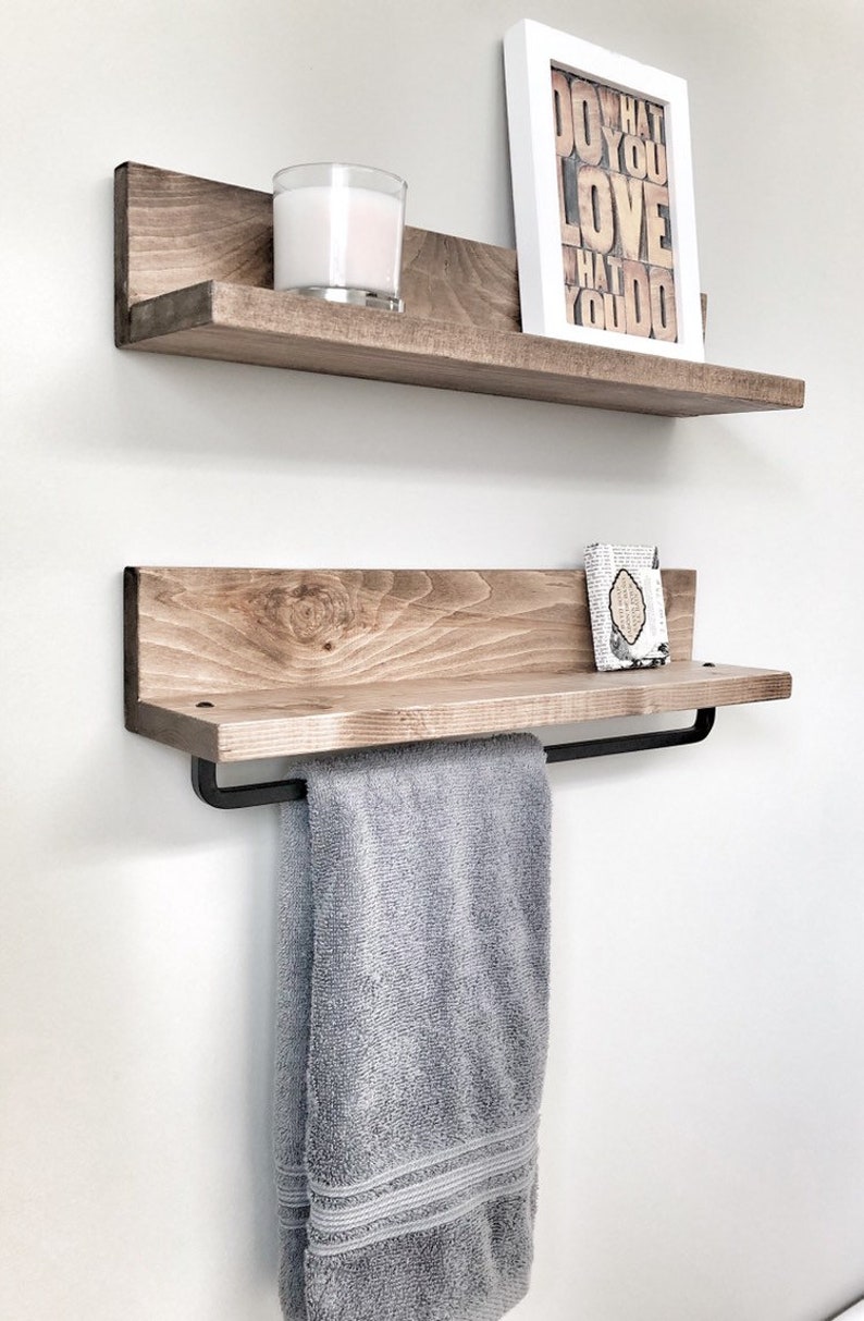 May include: Two wooden floating shelves with a light brown finish are mounted on a white wall. The top shelf holds a candle and a framed print with the text "DO WHAT YOU LOVE". The bottom shelf has a towel bar and a gray towel.