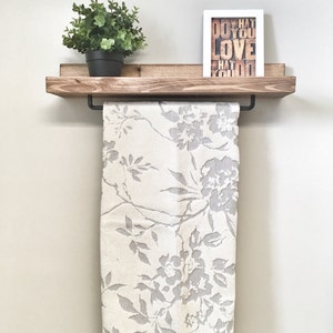 Op de afbeelding: Een houten plank met een zwarte handdoekstang, met een witte handdoek met bloemenpatroon. Een kleine potplant en een ingelijste print met de woorden "DO WHAT YOU LOVE" staan op de plank. De plank is aan een lichtgekleurde muur bevestigd.