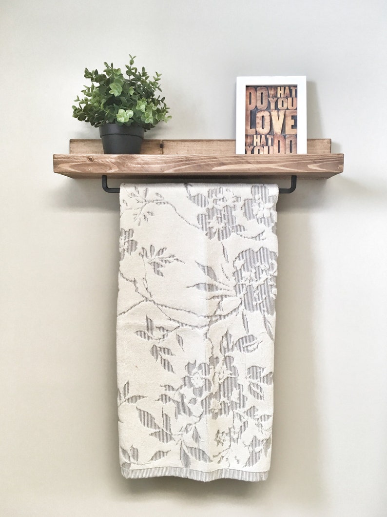 Op de afbeelding: Een houten plank met een zwarte handdoekstang, met een witte handdoek met bloemenpatroon. Een kleine potplant en een ingelijste print met de woorden "DO WHAT YOU LOVE" staan op de plank. De plank is aan een lichtgekleurde muur bevestigd.