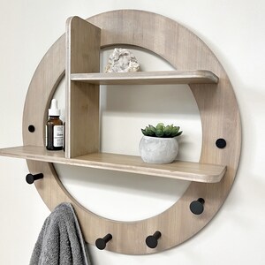 May include: A round wooden wall shelf with two shelves and five hooks. The shelf is made of light wood and has a natural finish. The hooks are black metal. There is a small plant in a pot on the top shelf, a crystal on the middle shelf, and a towel hanging on one of the hooks.