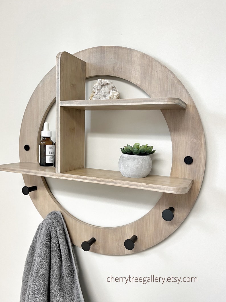 May include: A round wooden wall shelf with two shelves and five hooks. The shelf is made of light wood and has a natural finish. The hooks are black metal. There is a small plant in a pot on the top shelf, a crystal on the middle shelf, and a towel hanging on one of the hooks.