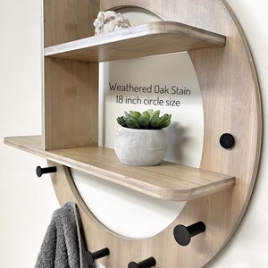 May include: A weathered oak stain wood wall shelf with three shelves and four black metal hooks. The shelf is in the shape of a circle with a diameter of 18 inches. The shelf is decorated with a small potted succulent plant and a crystal.