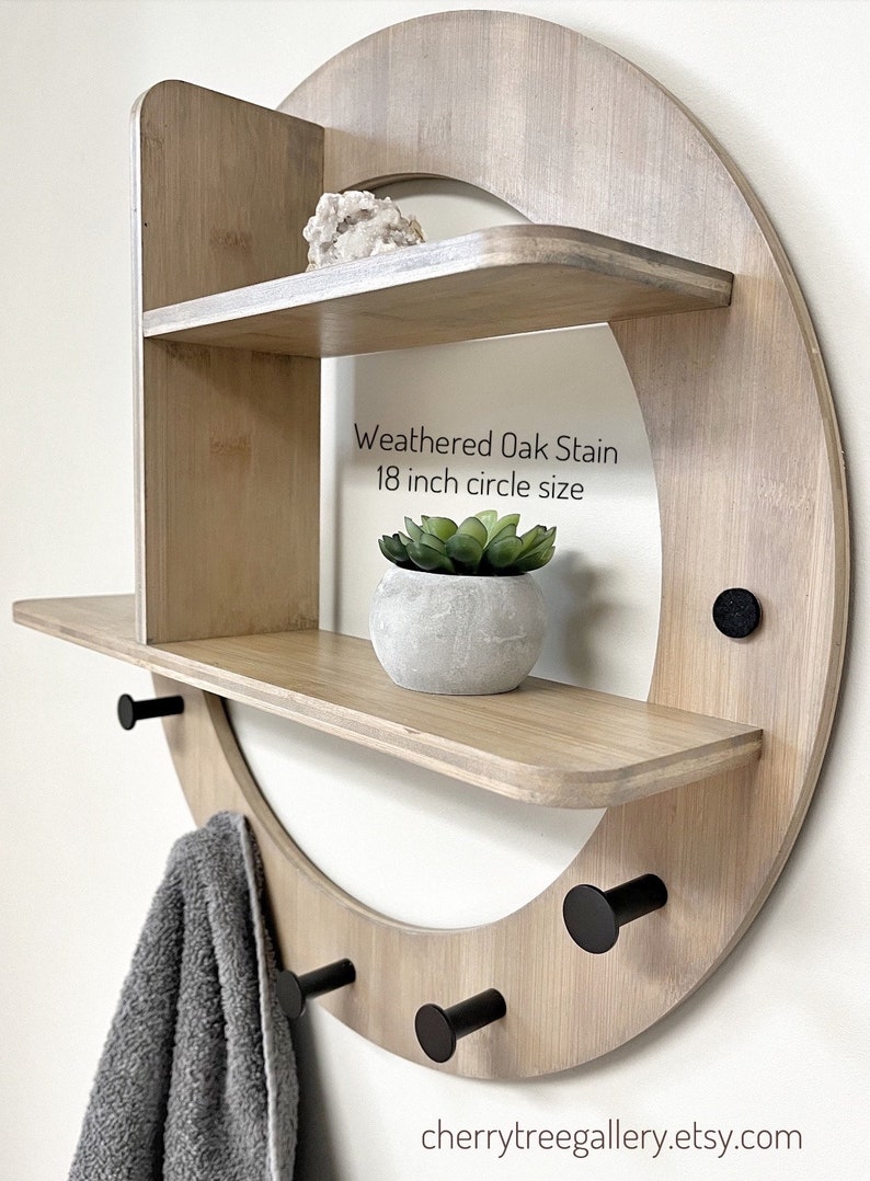 May include: A weathered oak stain wood wall shelf with three shelves and four black metal hooks. The shelf is in the shape of a circle with a diameter of 18 inches. The shelf is decorated with a small potted succulent plant and a crystal.