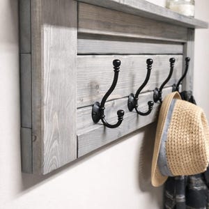 May include: A gray wooden wall-mounted coat rack with four black metal hooks. The rack has a shelf on top and is holding a straw hat and a scarf. The wood has a distressed finish.