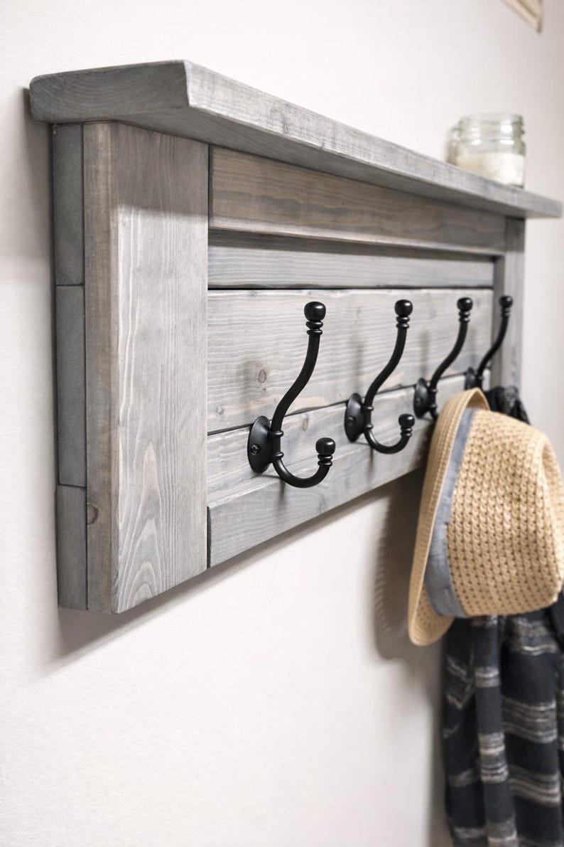 May include: A gray wooden wall-mounted coat rack with four black metal hooks. The rack has a shelf on top and is holding a straw hat and a scarf. The wood has a distressed finish.
