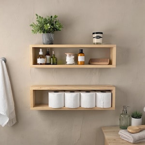 May include: Two wooden floating shelves mounted on a beige wall. The top shelf holds a potted plant, bottles, a jar, and a candle. The bottom shelf displays rolls of toilet paper, suggesting a bathroom setting.
