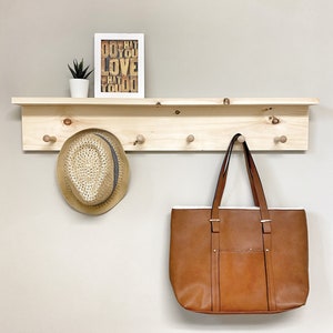 May include: A wooden shelf with five hooks and a small plant in a white pot. A brown leather tote bag hangs on one of the hooks. A straw hat with a grey band sits on another hook. A framed print with the text "Do what you love what you do" sits on the shelf.