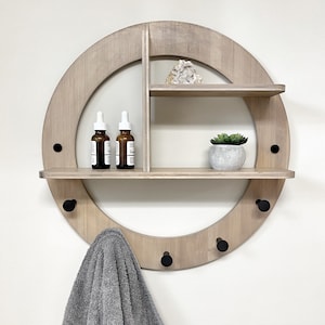 May include: A round wooden wall shelf with three hooks and two shelves. The shelf has a light wood finish and black metal hooks. A towel is hanging on one of the hooks. There are three small bottles on the top shelf and a small potted plant on the bottom shelf.