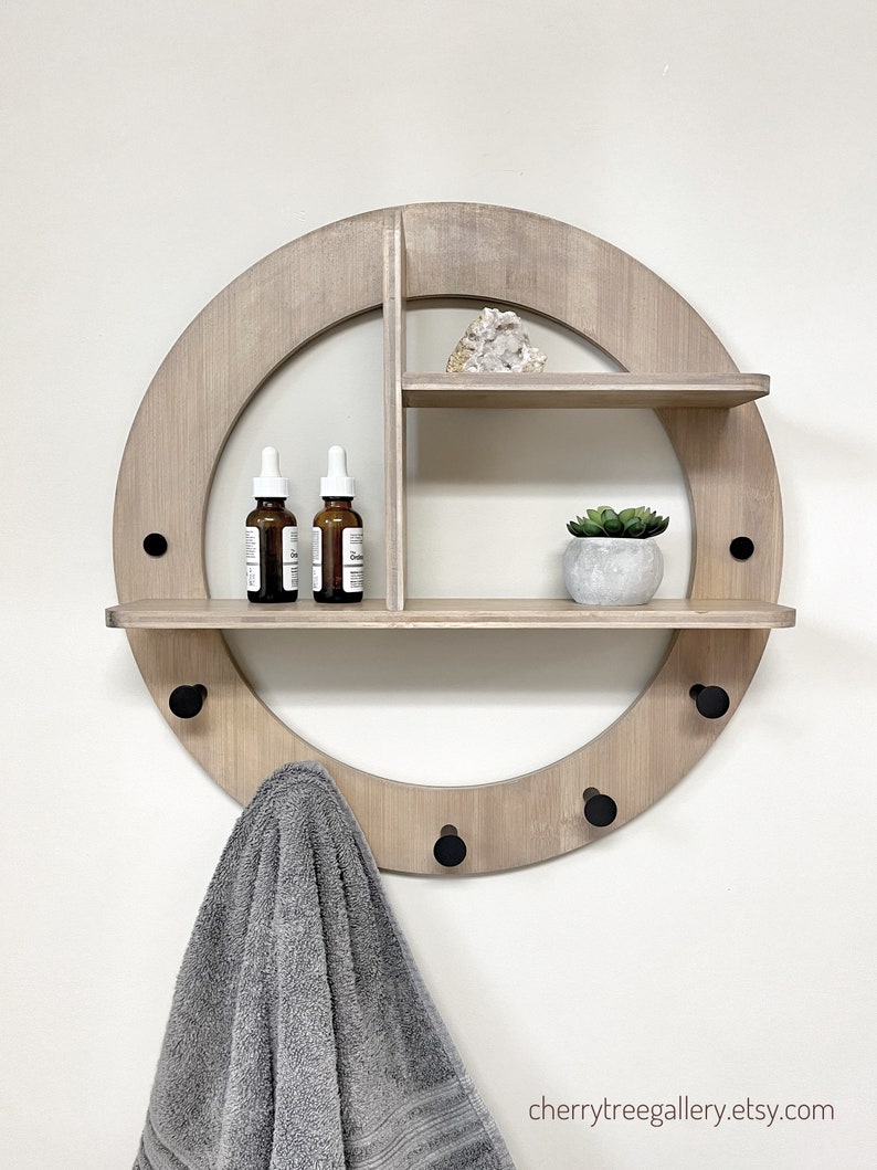 May include: A round wooden wall shelf with three hooks and two shelves. The shelf has a light wood finish and black metal hooks. A towel is hanging on one of the hooks. There are three small bottles on the top shelf and a small potted plant on the bottom shelf.