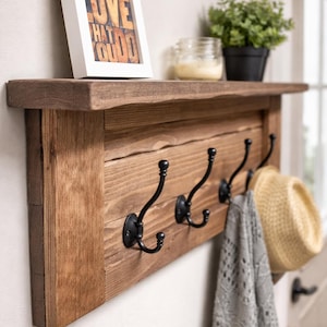 May include: A rustic wooden wall shelf with four black metal hooks. A framed print with the text "DO WHAT YOU LOVE" sits on the shelf, along with a small potted plant and a jar. A hat and a gray scarf hang from the hooks.