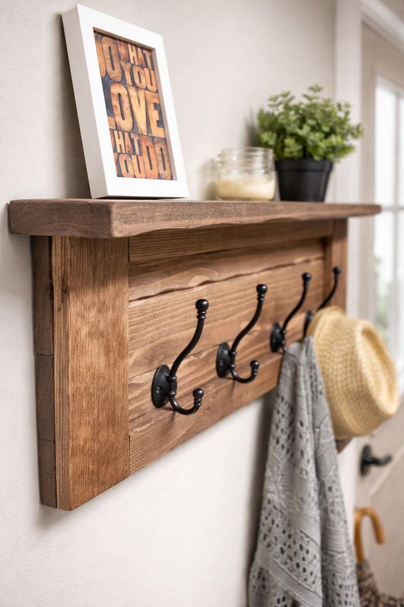 May include: A rustic wooden wall shelf with four black metal hooks. A framed print with the text "DO WHAT YOU LOVE" sits on the shelf, along with a small potted plant and a jar. A hat and a gray scarf hang from the hooks.