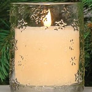 May include: A clear glass candle holder with a lit, cream-colored candle inside. The glass is decorated with a snowflake pattern around the top and bottom. The candle flame is visible, and the background is blurred.