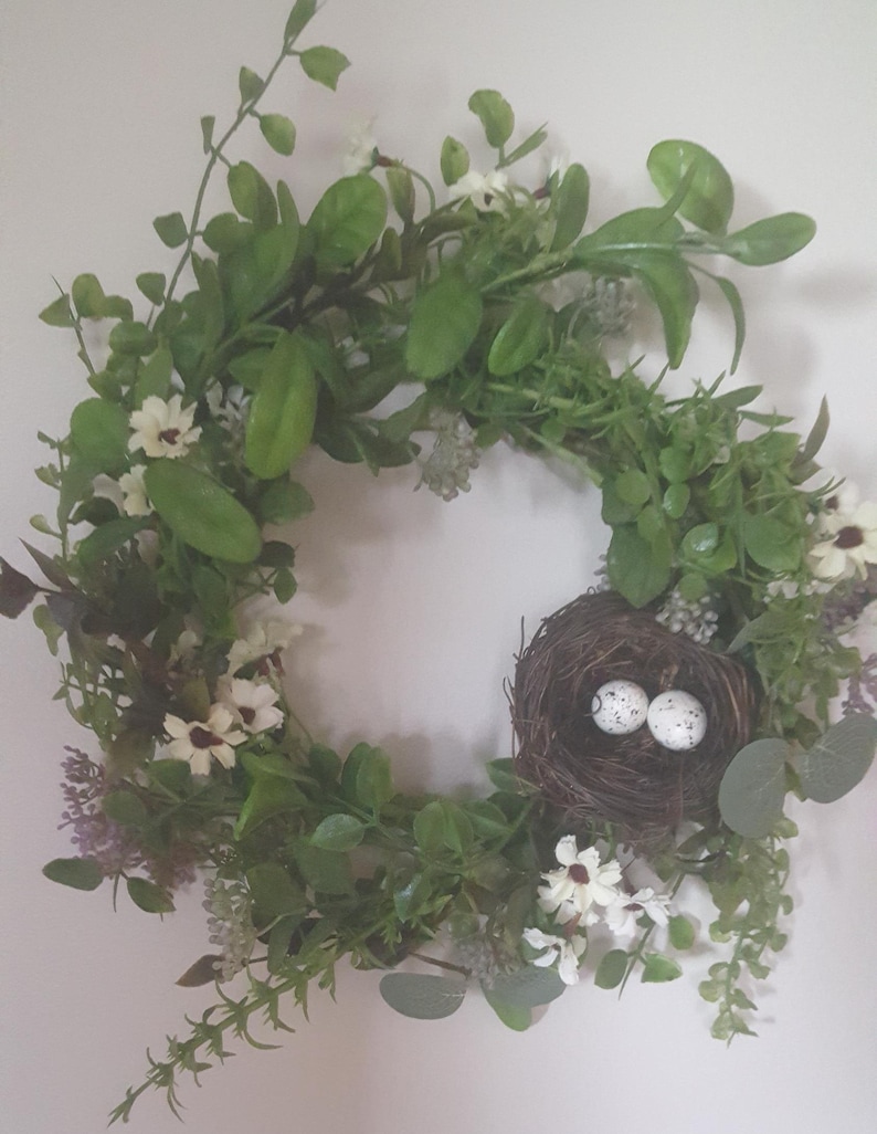 K&ouml;nnte beinhalten: Ein kreisf&ouml;rmiger Kranz aus gr&uuml;nen Bl&auml;ttern, kleinen wei&szlig;en Bl&uuml;ten und einem braunen Vogelnest mit zwei gesprenkelten Eiern. Der Kranz ist ein dekoratives Element, wahrscheinlich f&uuml;r die saisonale oder Heimdekoration.