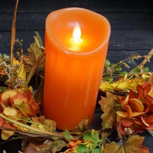 May include: A cylindrical orange LED pillar candle with a glowing white flame tip, surrounded by a decorative arrangement of artificial autumn-colored leaves, small yellow flowers, and dried grasses in shades of orange, yellow, and green.