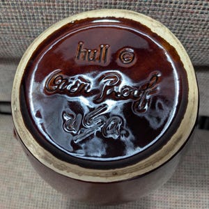 Pode incluir: Vista inferior de uma caneca de cerâmica castanha com uma borda creme. A base apresenta texto em relevo que diz "hull", "Oven Proof" e "USA". A caneca tem um acabamento brilhante.