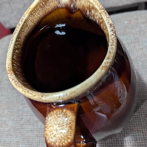 Puede incluir: Una jarra de cerámica marrón vintage con un acabado brillante. La jarra tiene una abertura ancha, un asa curva y un borde de color más claro. El interior es marrón oscuro y el exterior tiene un tono rico y cálido. Una pieza clásica para servir bebidas.