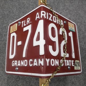 May include: A maroon Arizona license plate with white lettering and numbers, reading "D-74921" and "GRAND CANYON STATE." The plate is attached to a wooden post with a gold chain.