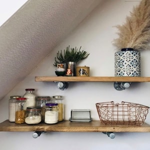 May include: Two wooden shelves with metal brackets, mounted on a white wall. The shelves are filled with various glass jars, a ceramic vase with dried pampas grass, and other decorative items.