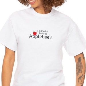 Puede incluir: Camiseta blanca con una manzana roja y el texto "Firmé un DNR en Applebee's"