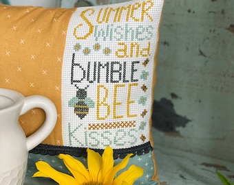 May include: A yellow and blue pillow with cross-stitch embroidery that reads "Summer Wishes and Bumble Bee Kisses". A sunflower and white daisies are in the foreground.