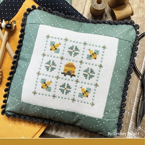 May include: A square, cross-stitched pillow with a bee and honeycomb design. The pillow has a green border with white polka dots and a black pom-pom trim. The text "August Nine Patch" and "Primrose Cottage Stitches" are visible.