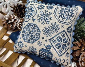 May include: A white and blue cross-stitch pillow with a snowflake design. The pillow is in a wooden crate with blue fabric and pine branches. The text "Winter Quaker" and "Primrose Cottage" are visible.