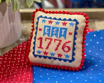 May include: A small, square, beige pillow with red pom-poms around the edge. The pillow is embroidered with blue and red stars, blue banners, and the text "1776".