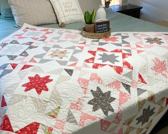 May include: A white quilt with a star pattern in shades of pink, red, and gray. The quilt is on a bed with a green headboard and pillows with the text "Let's Stay Home".