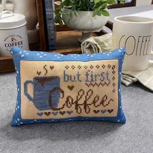 以下が含まれることがあります： コーヒーカップから蒸気が出ているスタイリッシュな画像と「but first, coffee」というテキストが書かれた青と茶色のクロスステッチのクッションです。