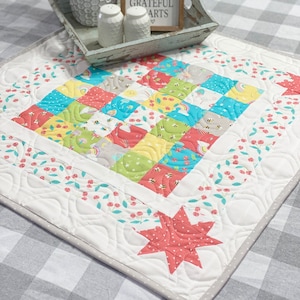 May include: A white quilted table runner with a patchwork design featuring a variety of colorful fabrics with patterns of bees, stars, rainbows, and cherries. The runner has a scalloped edge and a large red star in the bottom corner. The text "Gather here with grateful hearts" is visible on a wooden sign in the background.