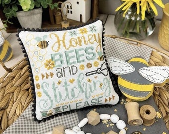 Puede incluir: Una almohada de punto de cruz con el texto "Honey Bees and Stitching Please" en hilo amarillo, verde y gris. La almohada está rodeada de una cesta de mimbre, cuentas de madera y una figurita de abeja de madera.