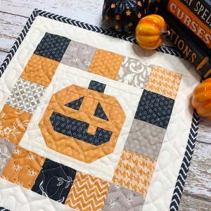 Pode incluir: Um pequeno corredor de mesa acolchoado com tema de Halloween com um design de patchwork que apresenta quadrados pretos, laranja e cinza. O quadrado central é uma abóbora laranja grande com uma cara preta.