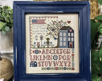 Könnte beinhalten: Ein gerahmter Kreuzstich-Sampler mit einem blauen Rahmen und einem weißen Passepartout. Der Sampler zeigt ein Haus, einen Baum, Vögel und den Text "My country tis of Thee sweet land of Liberty". Der Sampler enthält auch das Alphabet in Rot, Weiß und Blau.