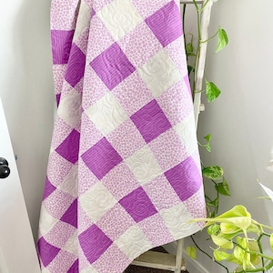 May include: A purple and white checkered quilt with a floral pattern. The quilt is folded and draped over a white wooden ladder.