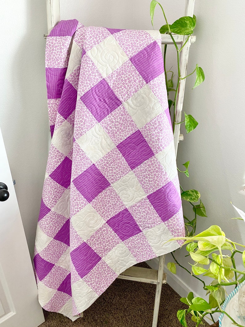 May include: A purple and white checkered quilt with a floral pattern. The quilt is folded and draped over a white wooden ladder.