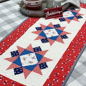 May include: A red, white, and blue quilted table runner with a star design. The runner has a white background with red and blue stars and anchors. The text "Proud to be an American" is on a red sign in the background.